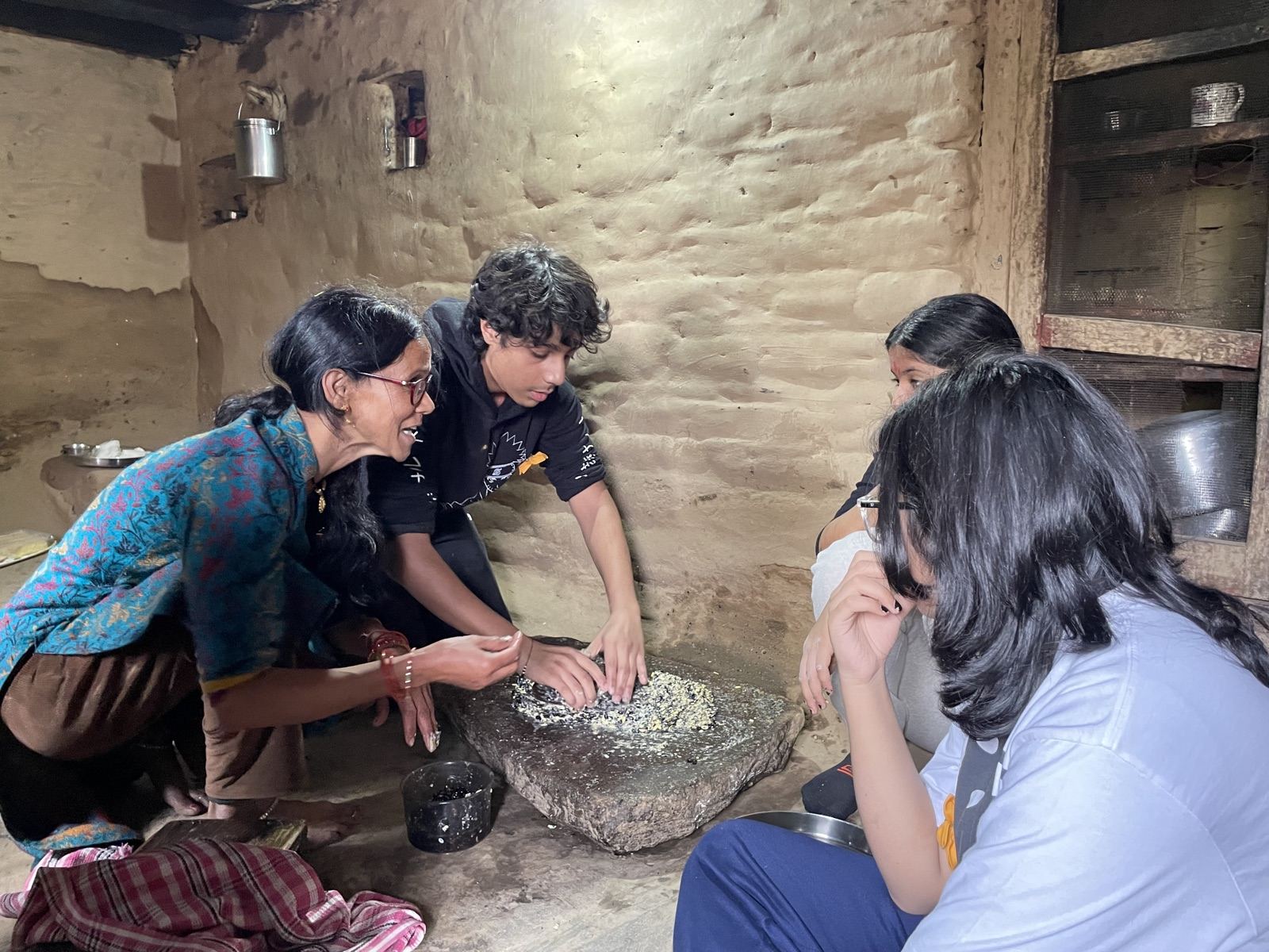 Learners grinding grain on a stone in a village kitchen at Majkhali